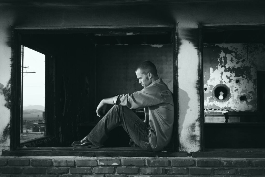 Ongelukkig leven. Man zit met zijn rug tegen de muur in een verlaten pand. Zwart-wit foto, met een sobere uitstraling. 