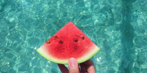 hand holding a slice of watermelon with blue swimming pool water in the background